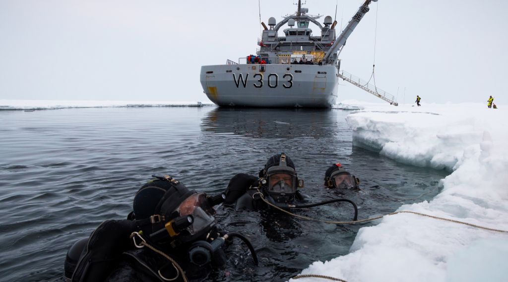 Tre dykkere i havet, ved iskanten. I bakgrunnen ser vi kystvaktens isbryter. 