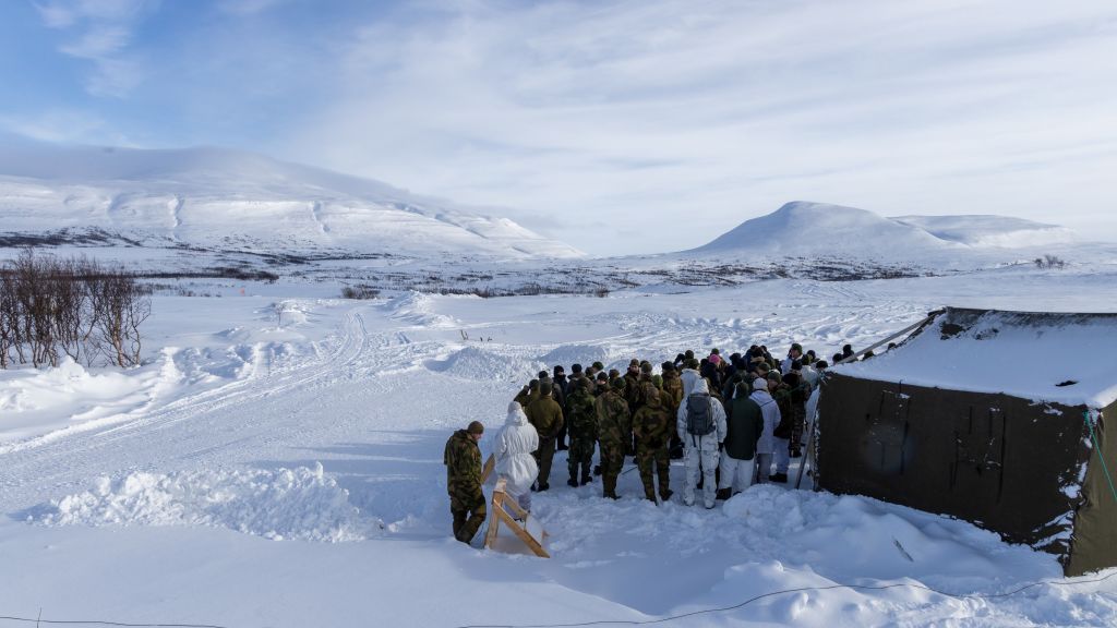 En gruppe mennesker i uniform ved et telt. Blå himmel. Utsikt over snødekte fjell og vidde.