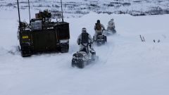 Tre soldater på snøscooter passerer en CV90 panservogn. Vinterlandskap. Finnmarksvidda. Fjell i bakgrunnen.