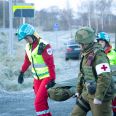 Tre personer bærer ei båre. To har sivil ambulanseuniform og en har militær uniform