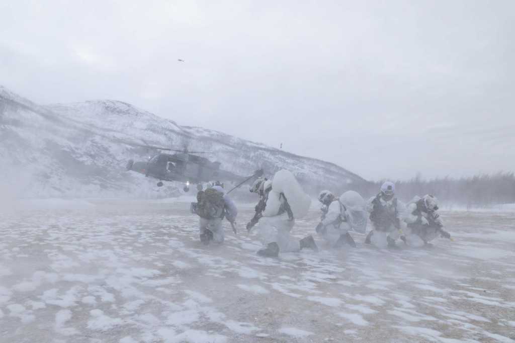 En gruppe soldater i hvite kamuflasjeuniformer venter på at et helikopter skal lande. Helikopteret pisker opp snøen rundt soldatene.