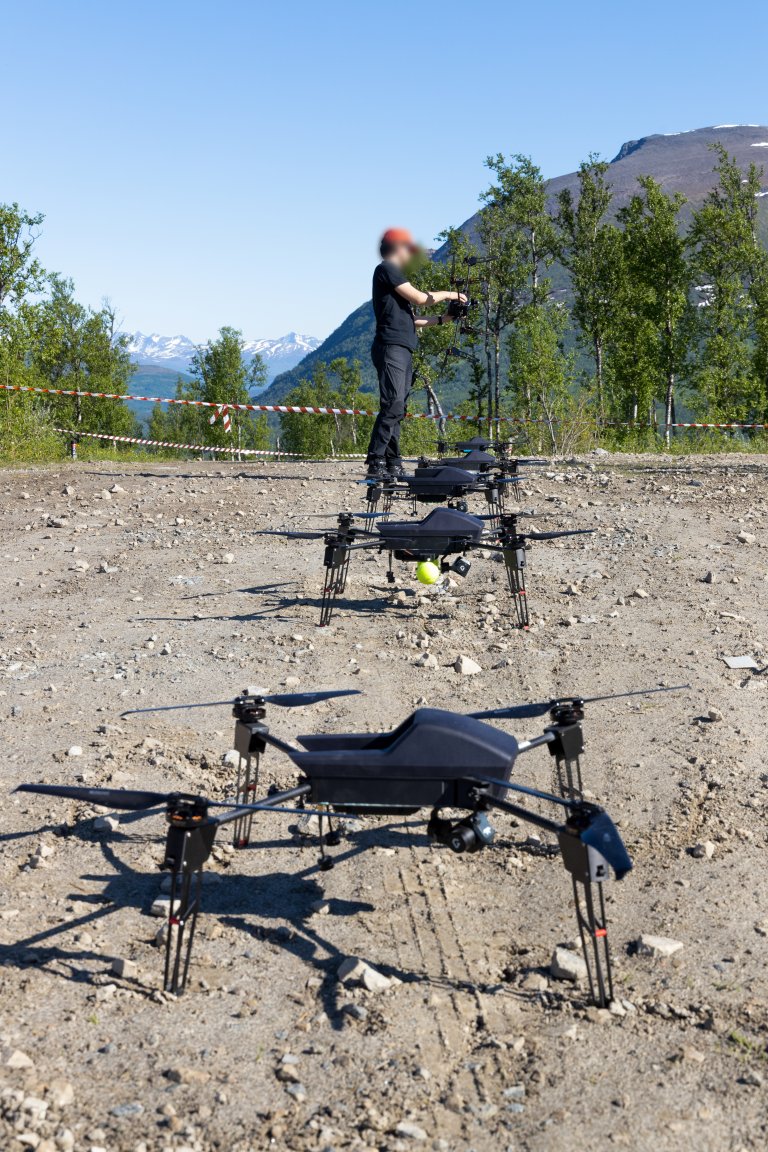En forsker gjør klar en dronesverm. Dronene står på rekke og rad på en grusplass, klare for takeoff. Sol, fjell. 