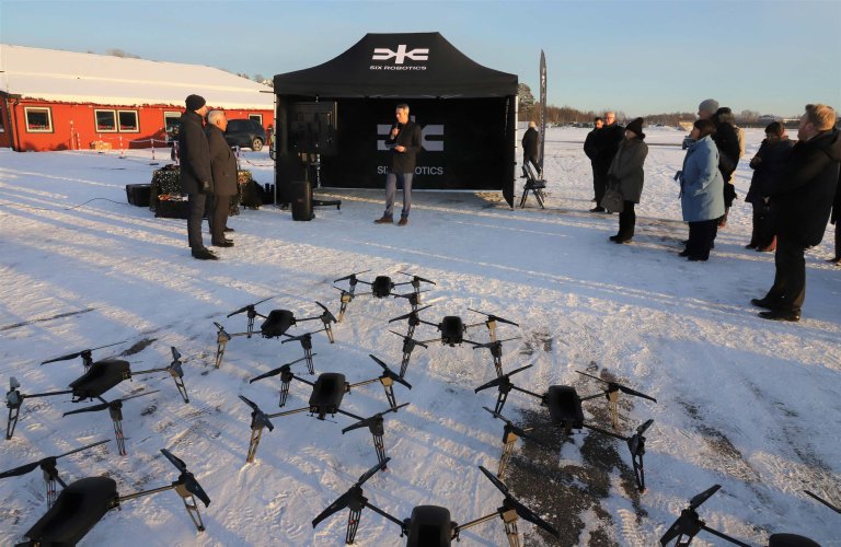 En rekke droner satt opp foran et telt. En person taler til en liten forsamling på en snødekt Kjeller flyplass.