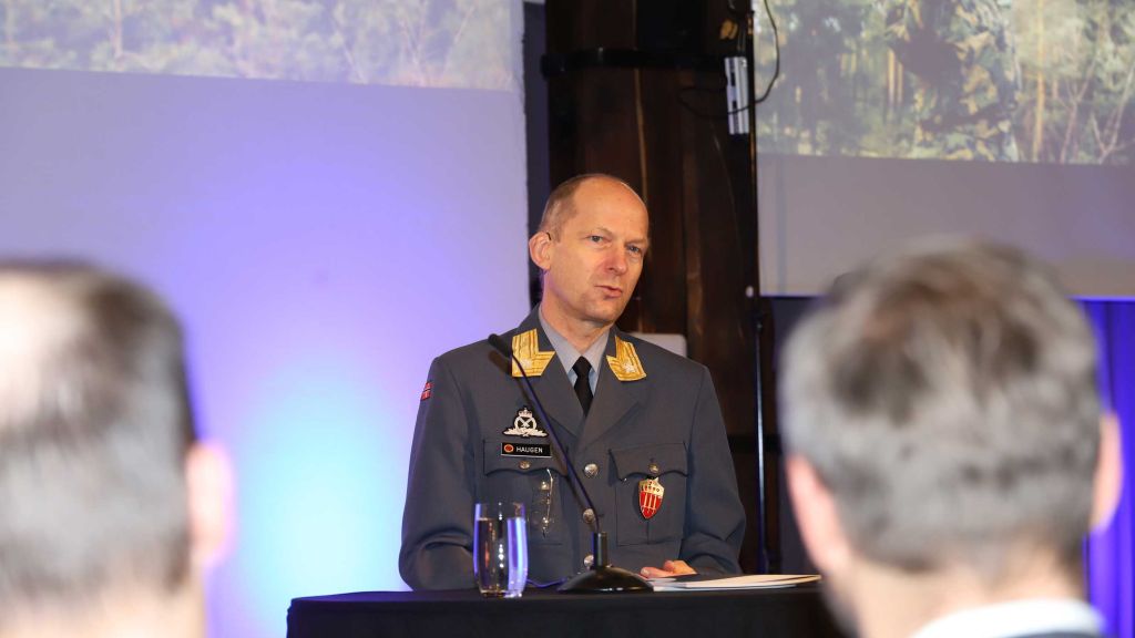 Mann i militæruniform på en talerstol. Publikum i forgrunnen.