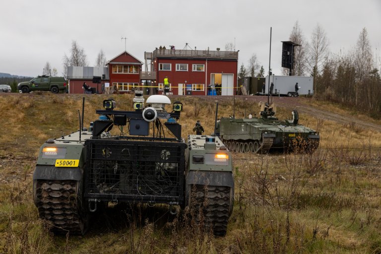 Et ubemannet kjøretøy og en CV90 panservogn foran FFIs arena for teknologisk eksperimentering. Høst. Grø himmel. Røde bygninger i bakgrunnen.