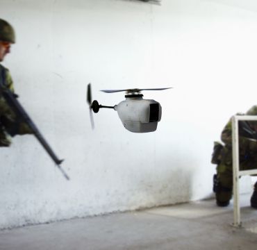 Liten drone i forgrunn, soldat med våpen i bakgrunn. Foto
