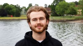 Ung mann i hettegenser står utendørs med havet i bakgrunnen. Ser rett i kamera og smiler