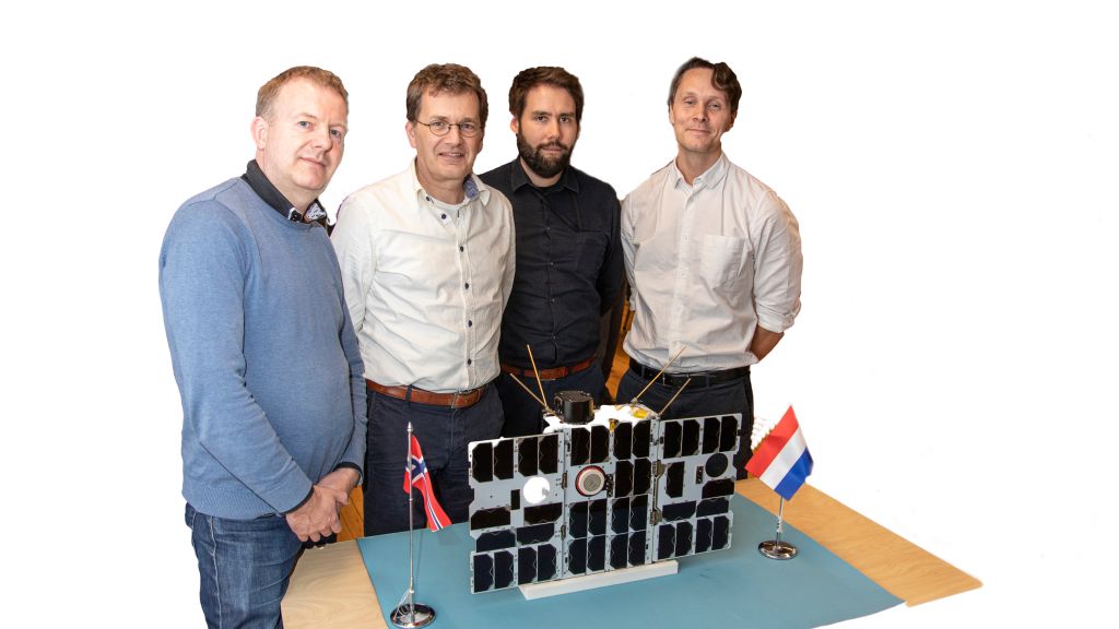 Fire menn står bak en mikrosatellitt. Norsk og nederlandsk flagg på bordet.