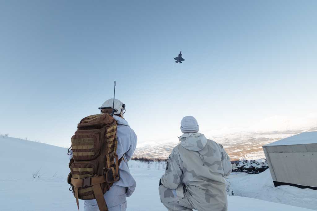 Bilde av to soldater i vinterkamuflasje med ryggen til kamera. Den ene har stor sekk med antenne og kommunikasjonsutstyr. Over dem flyr et F-35 kampfly. Vinter, snø og Nord-Norsk fjellandskap i bakggrunnen. 