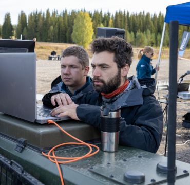 To menn ser på laptop mens de står utendørs på et militært øvingsfelt
