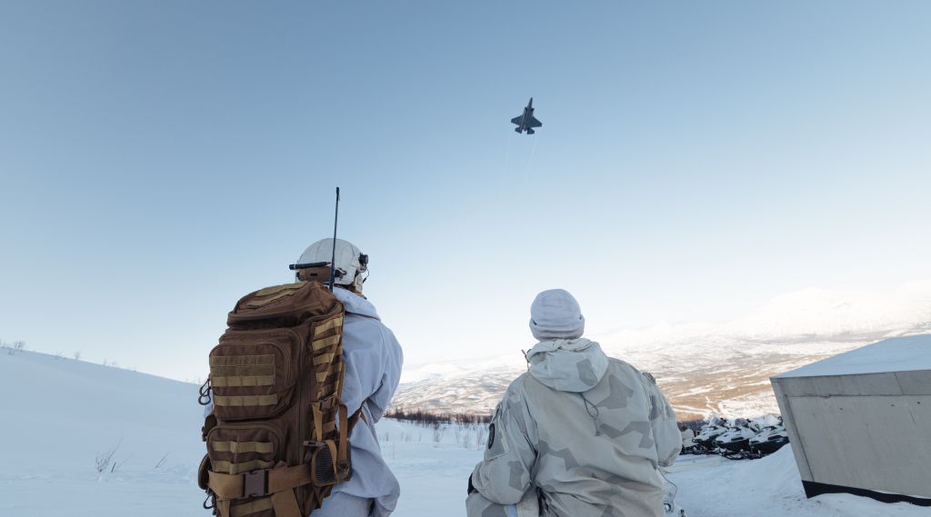 Bilde av to soldater i vinterkamuflasje med ryggen til kamera. Den ene har stor sekk med antenne og kommunikasjonsutstyr. Over dem flyr et F-35 kampfly. Vinter, snø og Nord-Norsk fjellandskap i bakggrunnen. 