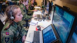 en ung kvinne i grønn militæruniform og norsk flagg på armen sitter med hodetelefoner og ser på flere dataskjermer