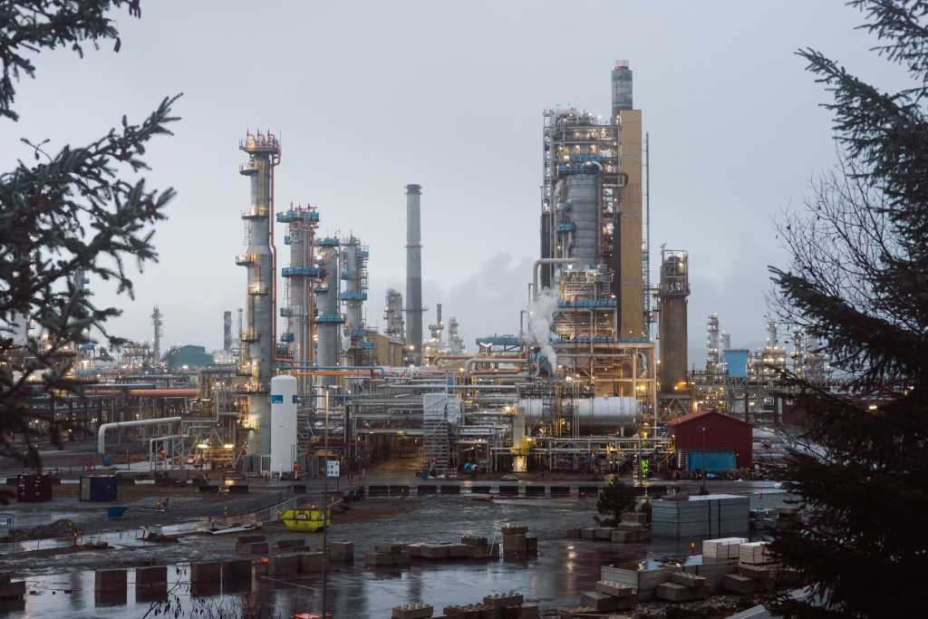 BIlde av oljeraffineri tatt i grått vintervær. Rør, tanker og lys. I forgrunnen to grantrær.