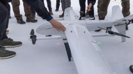 Flere personer samlet rundt en drone. Kun føtter og hender er synlig. 