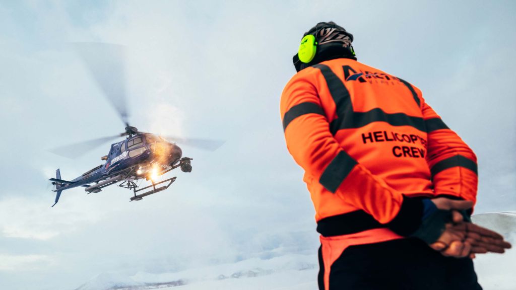 Et blått helikopter er på vei til å lande. En person i orange kjeldedress passer på.