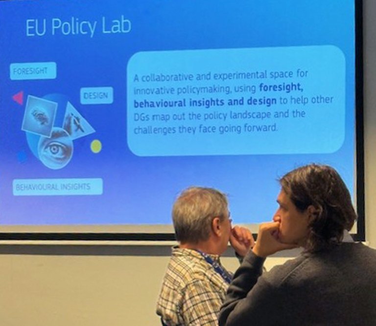 Bilde av ein presentasjon med overskrifta EU Policy Lab. To personar sit nede til høgre.