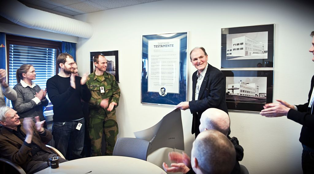 En eldre mann avduker en stor plakat i glass og ramme mens andre står rundt og klapper