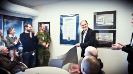 En eldre mann avduker en stor plakat i glass og ramme mens andre står rundt og klapper