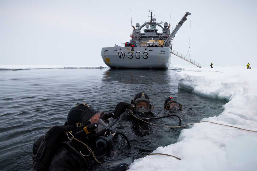 Tre dykkere i havet, ved iskanten. I bakgrunnen ser vi kystvaktens isbryter. 