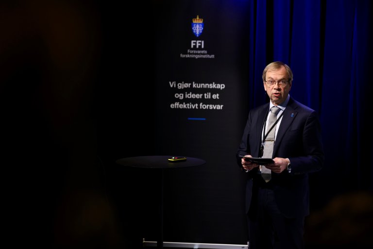 Mann i dress på scenen i Deichmansalen. I bakgrunnen en rollup med FFIs slagord. "Vi gjør kunnskap og ideer til et effektivt  forsvar".