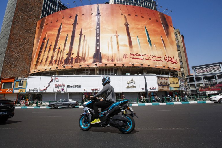 Mann på motorsykkel kjører forbi en gigantisk plakat av missiler midt i teheran. Blå himmel. Sommer. Trafikk.