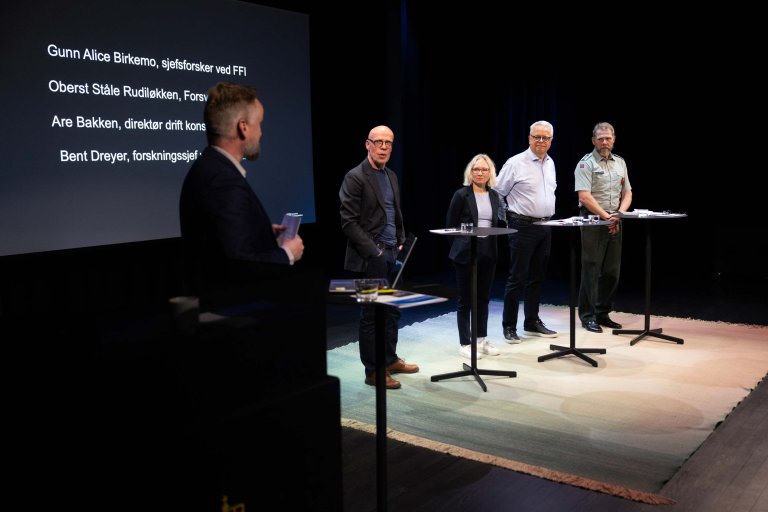 Fire menn og ei kvinne på scenen. En PowerPoint med navnene i bakgrunnen.
