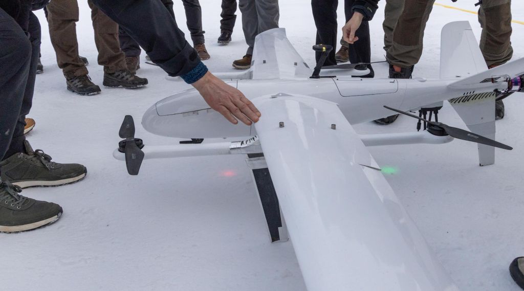 Flere personer samlet rundt en drone. Kun føtter og hender er synlig. 
