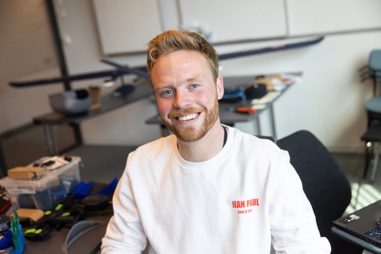 Student fotografert inne på et kontor. I bakgrunnen diverse verktøy og dronen han jobber med å bygge.