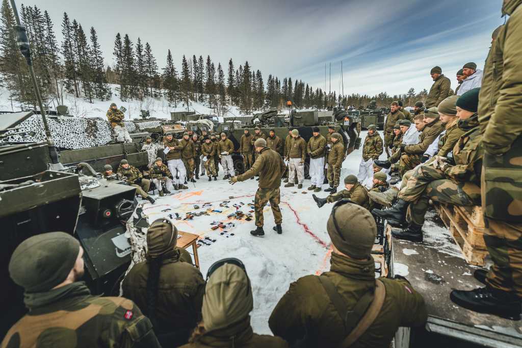 En gruppe soldater i sirkel rundt en offiser som tegner og forklarer. Utendørs. Snø og vinter. Militære kjøretøyer.