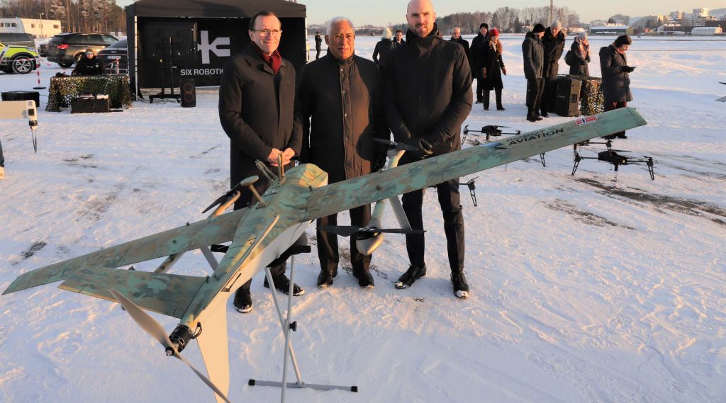 Tre menn poserer bak en drone på en snølagt flyplass.