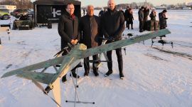 Tre menn poserer bak en drone på en snølagt flyplass.