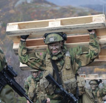 Soldat med full utrustning og kamuflasjemaling i ansiktet løper mens han bærer en europall på ryggen.