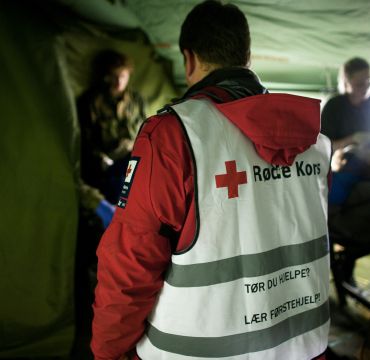 inne i et militært telt. vi ser tyggen på en med vest fra røde kors i fokus og annet militært personell rundt