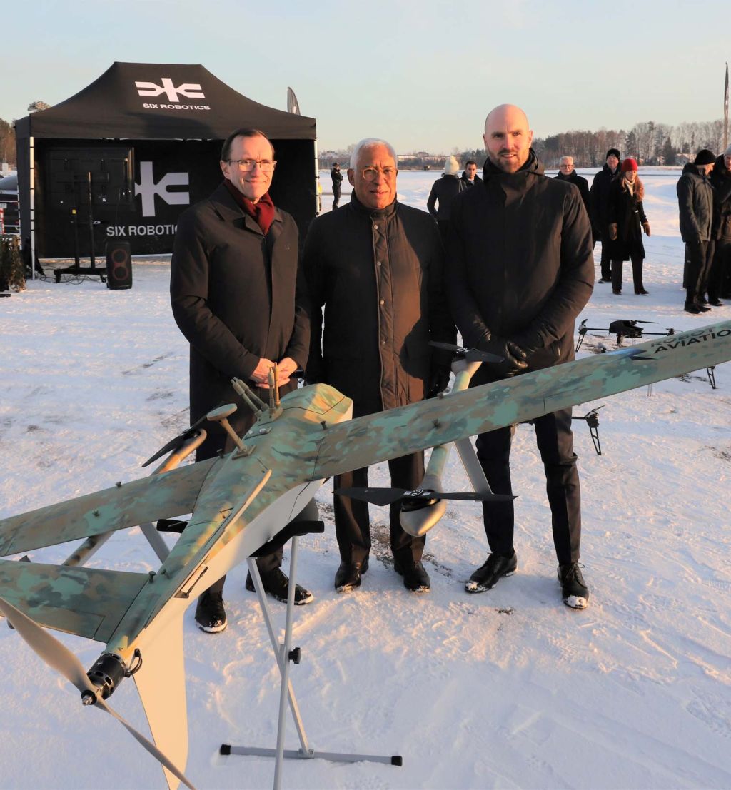 Tre menn poserer bak en drone på en snølagt flyplass.