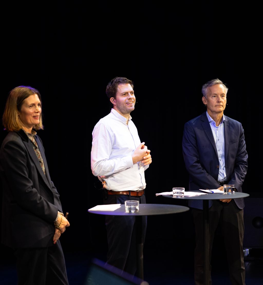 Tre personer på scenen i Deichmanssalen. Svart bakgrunn.
