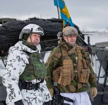 En finsk og en norsk soldat står i vinterbekledning ute foran et kjøretøy med svensk flagg