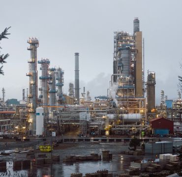 BIlde av oljeraffineri tatt i grått vintervær. Rør, tanker og lys. I forgrunnen to grantrær.