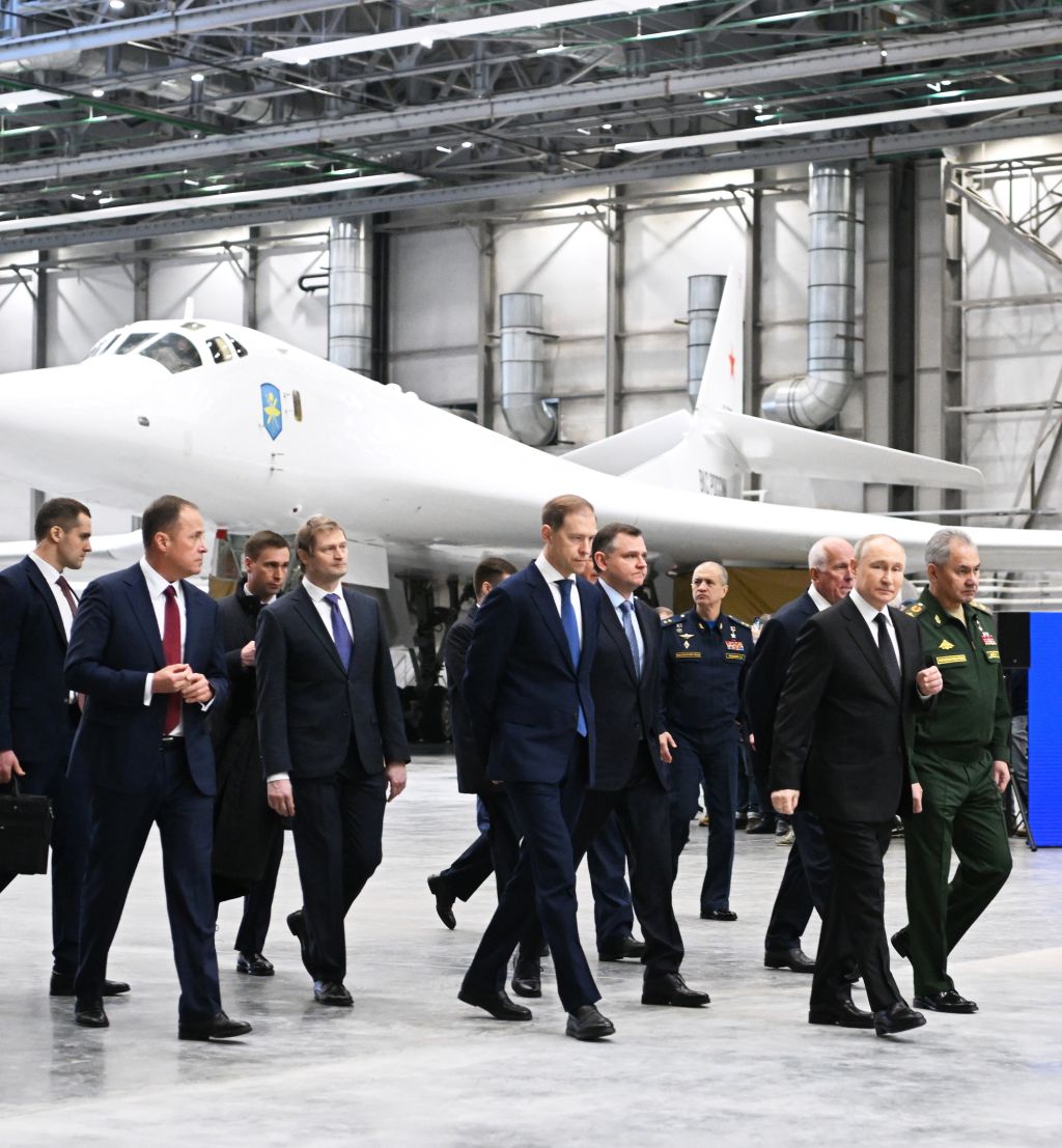 Menn i dress går rundt i en stor hangar med et moderne hvitt jagerfly i bakgrunnen