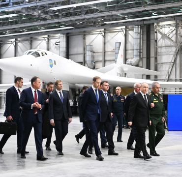 Menn i dress går rundt i en stor hangar med et moderne hvitt jagerfly i bakgrunnen