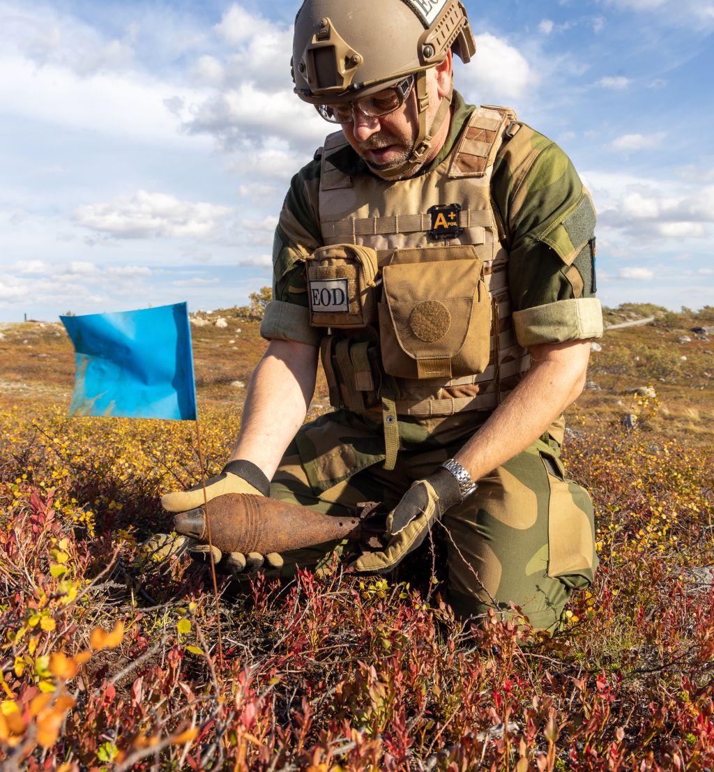 En soldat med splintvest, hjelpm og vernebruller sitter på kne i lyngen. I hendene holder han en rusten granat. Fjelllandskap og blå himmel.