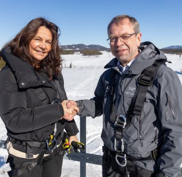 Direktør Birgitte Engebretsen i Telenor Norge og FFI-direktør Kenneth Ruud utveksler håndtrykk i toppen av en mast. Strålende blå himmel og norsk vinterlandskap i bakgrunnen.