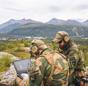 To militærkledde sitter med PC og radioutstyr på en fjellknaus i høstlandskap